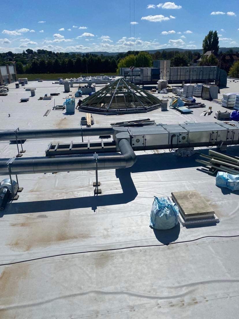 Widok z góry na płaski dach budynku z instalacją wentylacyjną, elementami konstrukcji świetlika i materiałami budowlanymi, w tle zabudowa miejska i drzewa.