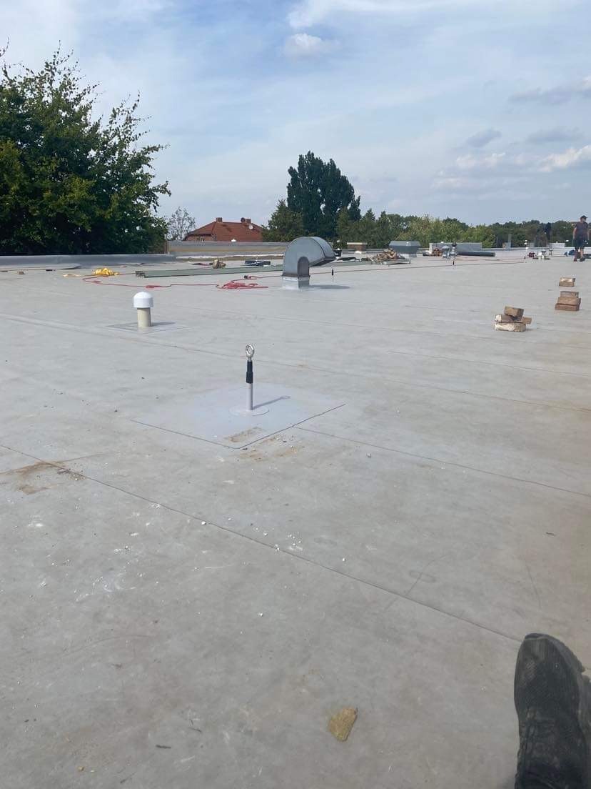 Płaski dach pokryty szarą membraną z widocznymi elementami wentylacyjnymi, mocowaniami i cegłami, prawdopodobnie w trakcie prac dekarskich, z butem w dolnym rogu kadru.