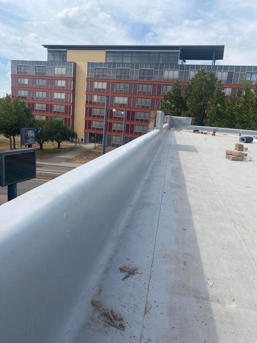 Płaski dach budynku wielorodzinnego z widoczną obróbką blacharską attyki, zabezpieczoną szarą blachą, z cegłami i narzędziami przygotowanymi do dalszych prac wykończeniowych.