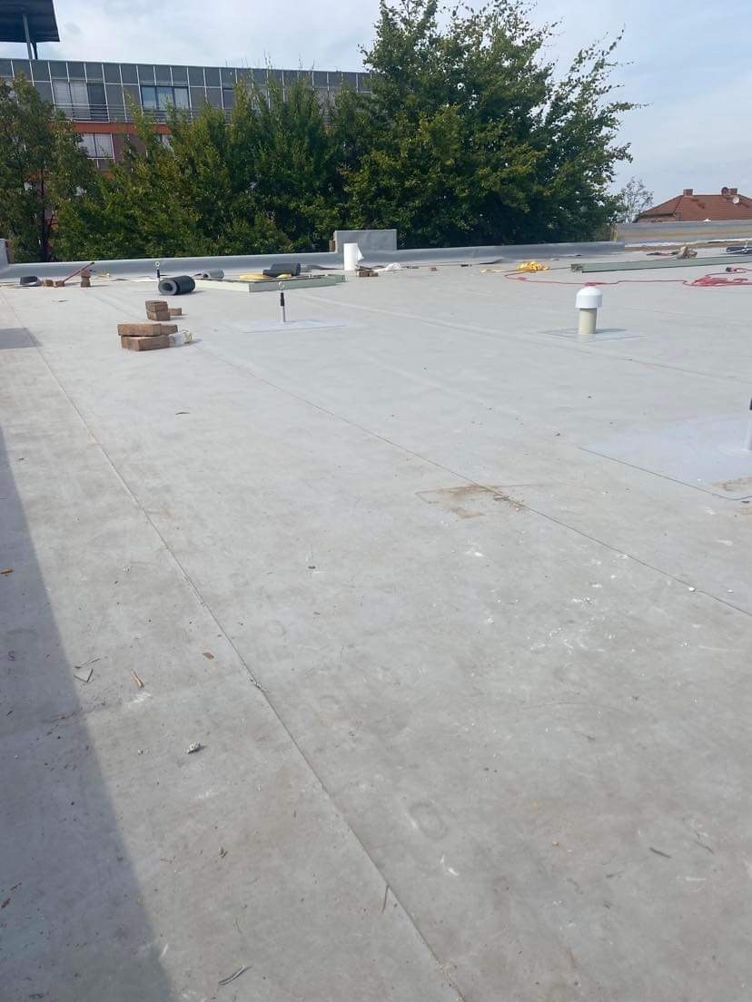 Płaski dach pokryty szarą membraną bitumiczną z widocznymi elementami instalacji, cegłami i narzędziami przygotowanymi do dalszych prac wykończeniowych, na tle zieleni i budynków.