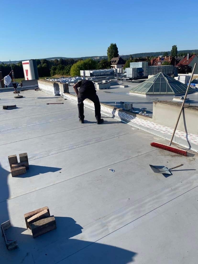 Pracownik w czarnym ubraniu pochylony na płaskim dachu pokrytym szarą membraną, cegły i narzędzia obok, widok na miasto w tle, słoneczny dzień.