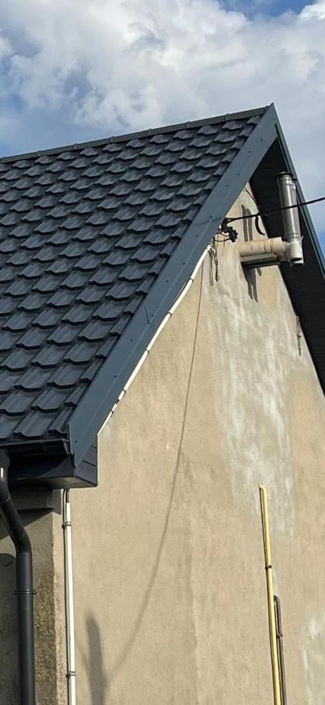 Fragment dachu z ciemną blachodachówką, szara obróbka blacharska, rynna spustowa i komin wentylacyjny na tle jasnego nieba i ściany budynku z widocznymi śladami zabrudzeń.