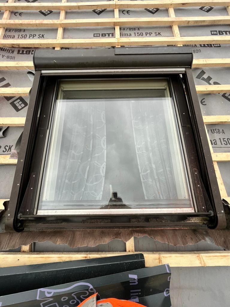 Okno dachowe z roletą zewnętrzną, widoczne elementy konstrukcji dachu i papa podkładowa z nadrukiem, fragmenty blachy na pierwszym planie.