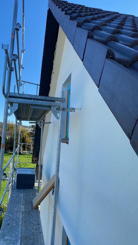 Fragment elewacji domu z nową, ciemną dachówką, widoczne rusztowanie i zabezpieczone okno podczas prac wykończeniowych.