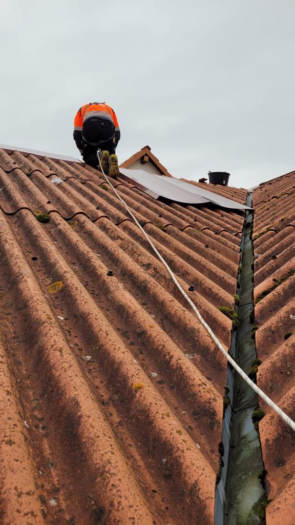 Pracownik w odblaskowej kurtce zabezpieczony liną na dachu z falistej dachówki ceramicznej z widocznymi zabrudzeniami i mchem, obok rynna z zanieczyszczeniami, pochmurne niebo.