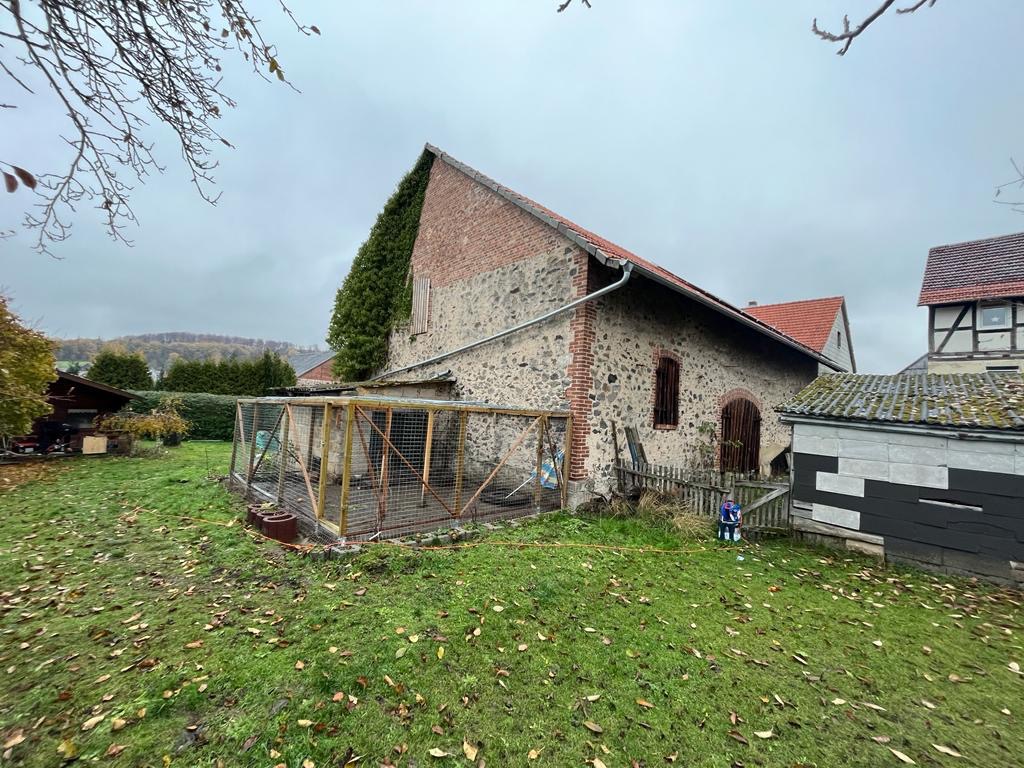 Kamienny budynek gospodarczy z ceglanym szczytem i rynną, widok z zewnątrz na tle pochmurnego nieba i jesiennego ogrodu z opadłymi liśćmi.