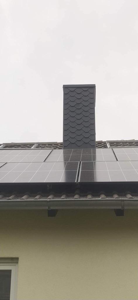 Dach pokryty panelami słonecznymi, obok komin wykończony łupkiem dachowym, widok z dołu na jasnym tle nieba.