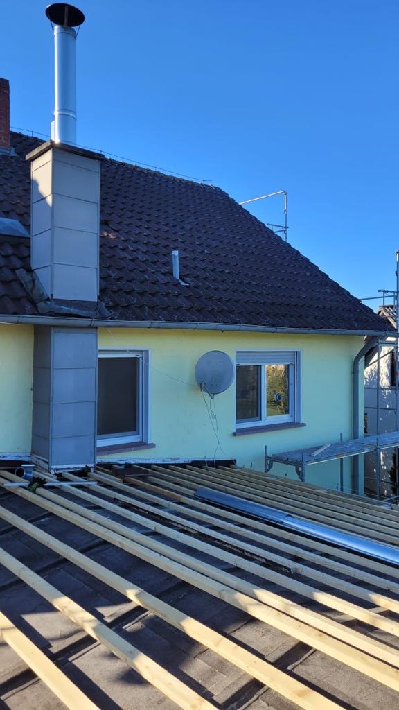 Dom w trakcie prac dekarskich: widoczne łaty na dachu, komin z blachy, antena satelitarna na elewacji.