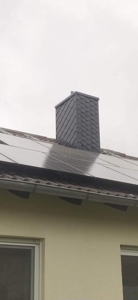 Szary komin obłożony łupkiem na dachu z panelami słonecznymi i brązową dachówką, widok z dołu na tle pochmurnego nieba.