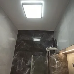 Nowoczesna kabina prysznicowa z ciemnymi płytkami imitującymi marmur, oświetlona kwadratowym panelem LED w suficie i dodatkowym oświetleniem punktowym nad prysznicem.