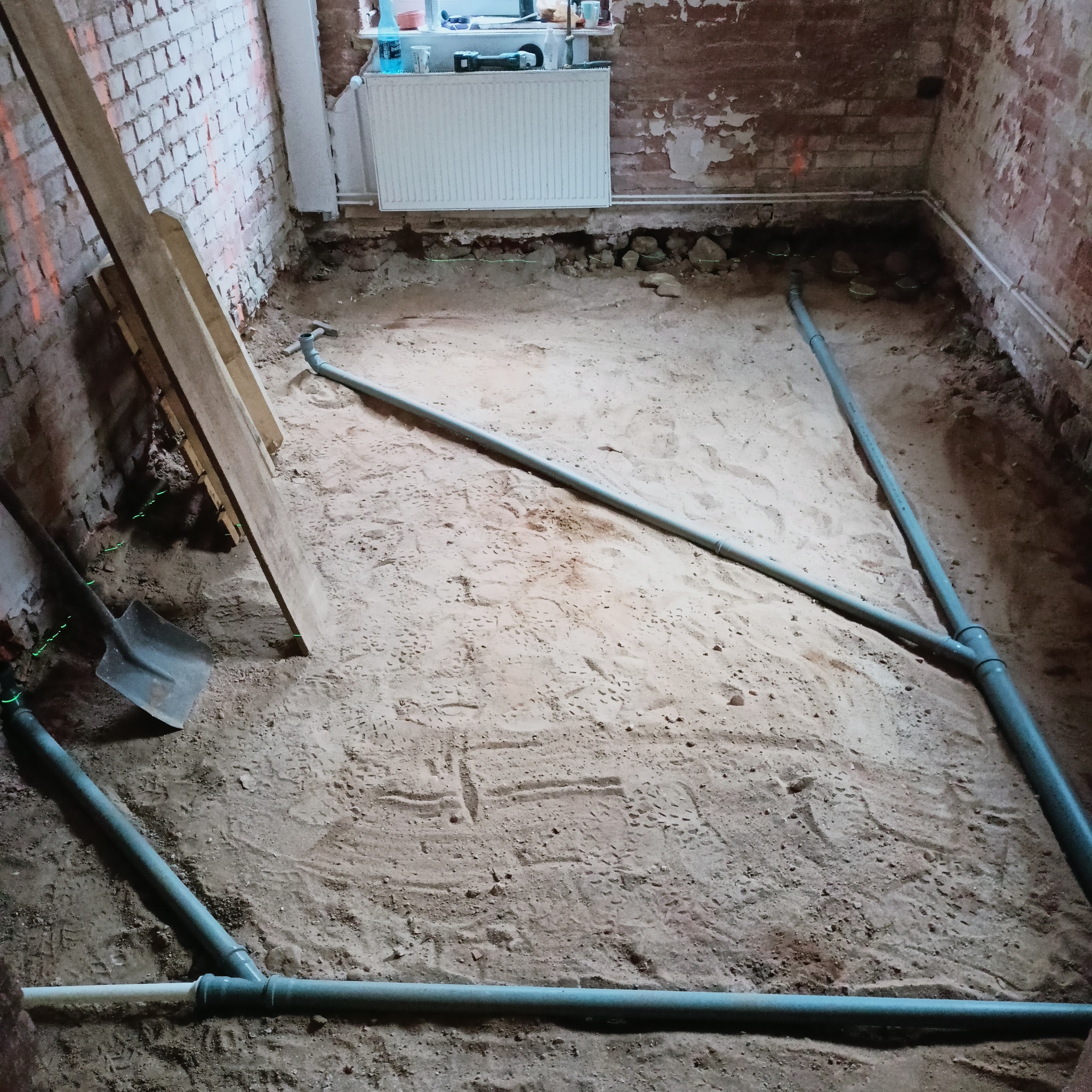 Przygotowanie podłoża w łazience przed remontem: widoczne odkryte cegły, piasek, rury kanalizacyjne, poziomica laserowa wyznaczająca poziom, deski i łopata.