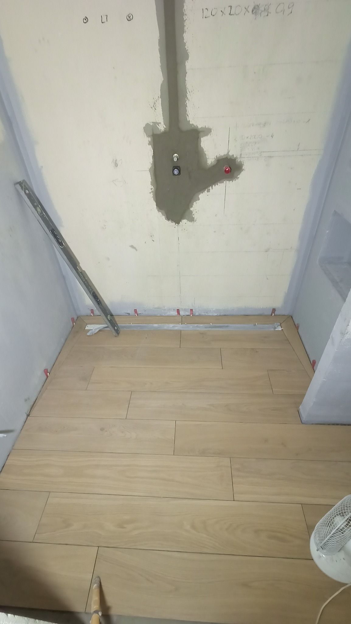 Podczas prac: Ułożona podłoga z płytek imitujących drewno w łazience, widoczne poziomice, krzyżyki dystansowe, przygotowane punkty instalacyjne na ścianie.