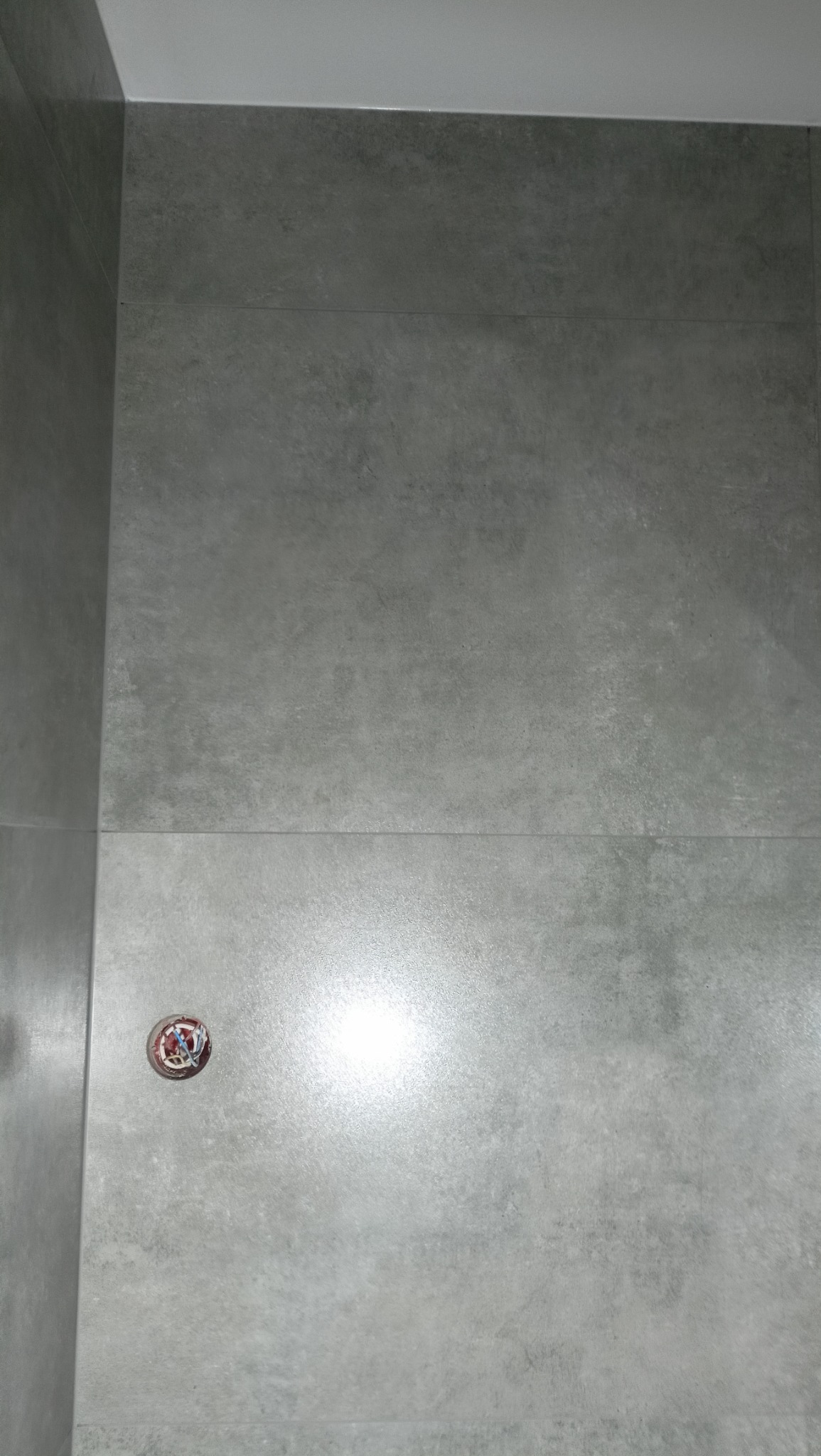 Ściana łazienki wyłożona szarymi płytkami imitującymi beton, z widocznym otworem na odpływ liniowy w podłodze, zabezpieczonym dekoracyjnym korkiem.