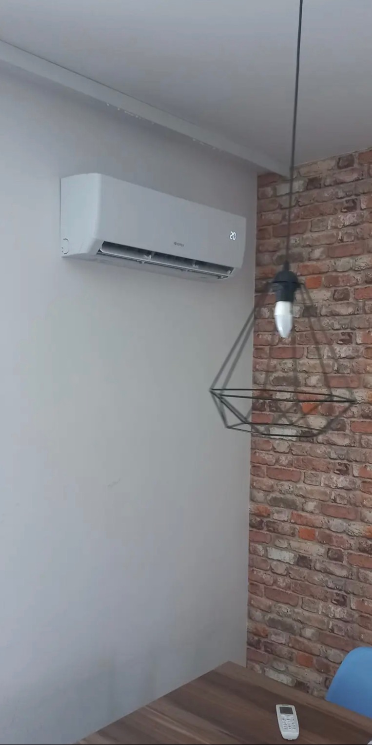 Biały klimatyzator ścienny marki Gree z wyświetlaną temperaturą 20 stopni Celsjusza, zamontowany na gładkiej, szarej ścianie obok ściany z dekoracyjnej cegły, w tle widoczna nowoczesna lampa...