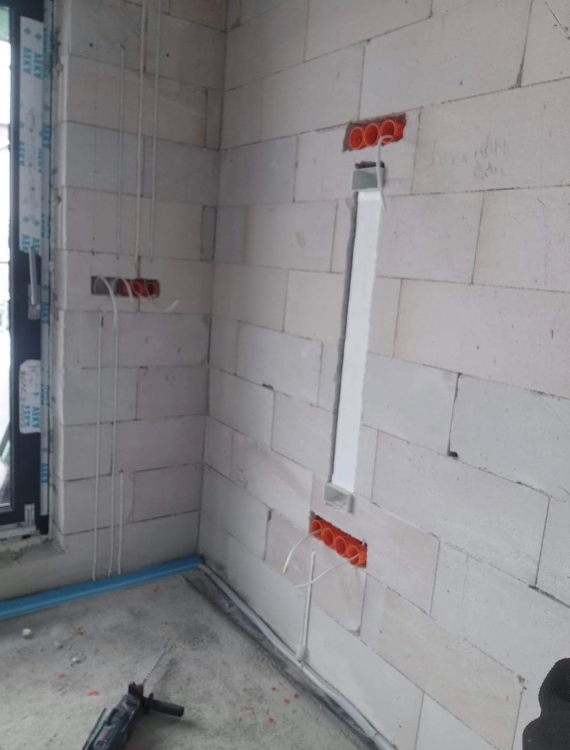 Ściana z betonu komórkowego w trakcie prac elektrycznych: widoczne puszki instalacyjne z przewodami, peszel pionowy, oraz przewody prowadzone wzdłuż ściany przy oknie, w tle wiertarka udarowa.