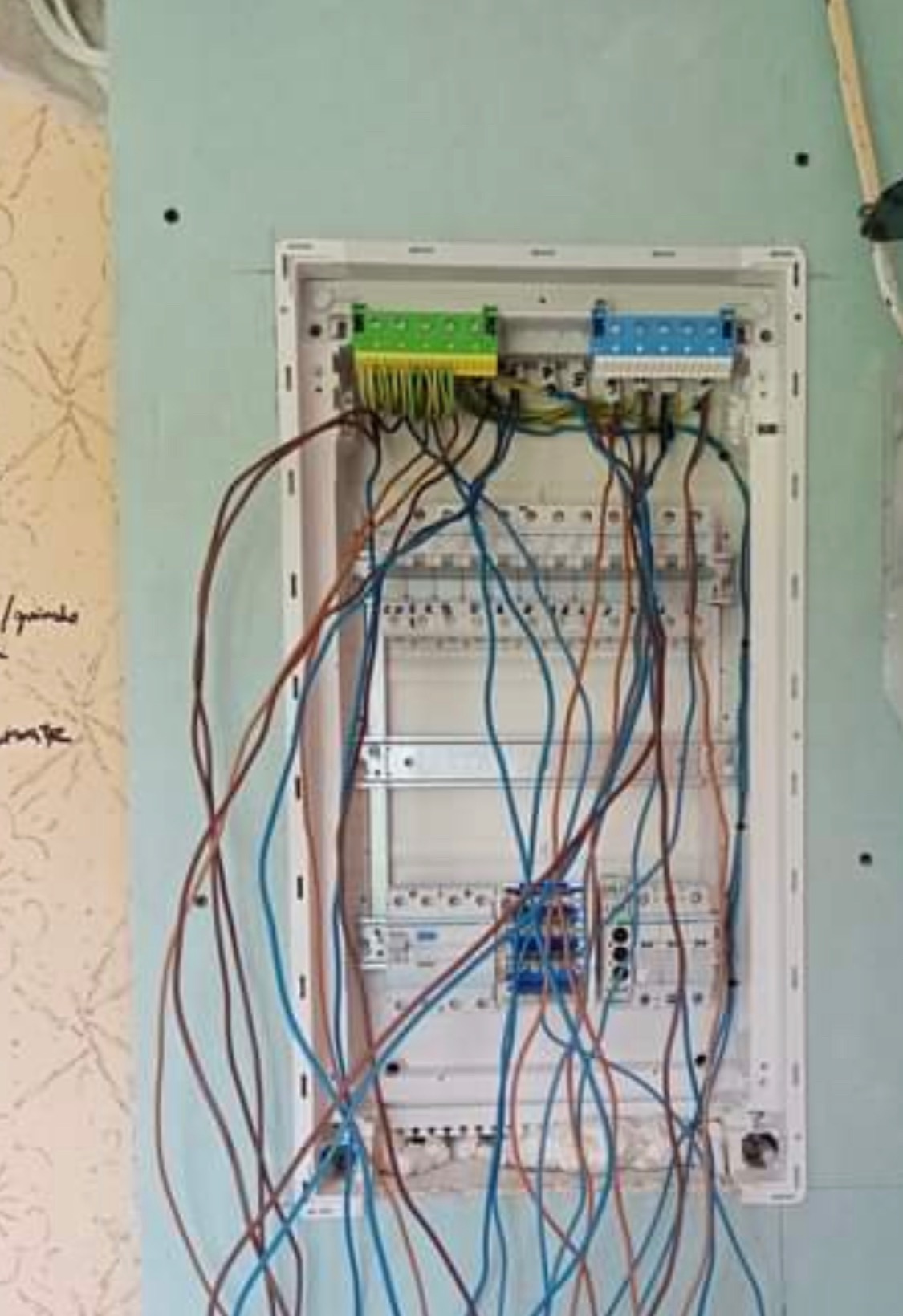 Rozdzielnica elektryczna w trakcie montażu, widoczne liczne przewody w kolorach niebieskim i brązowym, podłączone do zielono-żółtych i niebiesko-białych listew zaciskowych, zamontowana na ścianie...