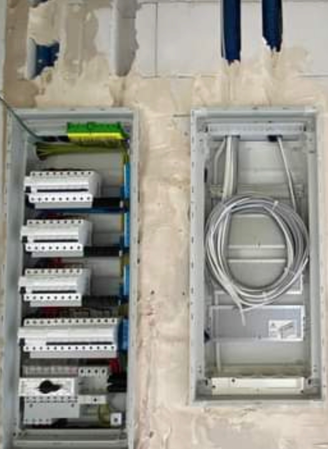 Dwie skrzynki elektryczne: jedna z zamontowanymi bezpiecznikami i okablowaniem, druga z zapasem przewodów, na tle surowej ściany z betonu komórkowego w trakcie prac wykończeniowych.