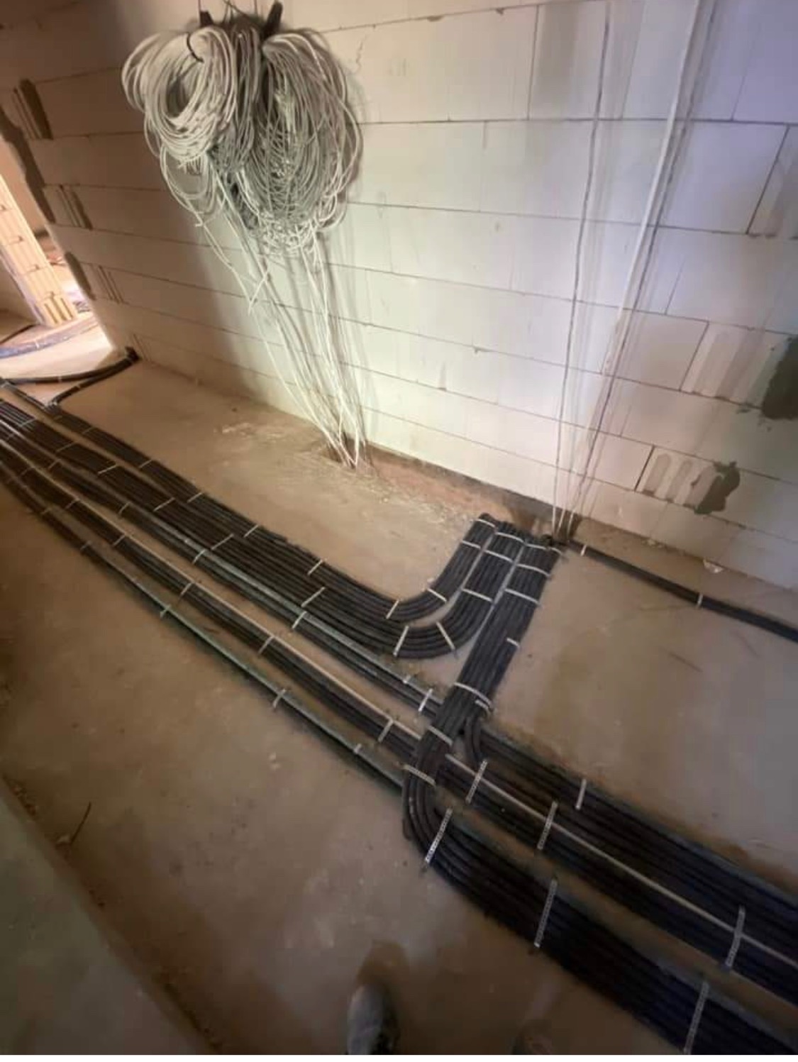 Układanie przewodów elektrycznych w rurach ochronnych na podłodze w surowym stanie; ze ściany zwisają białe kable. Widoczny fragment buta montera.