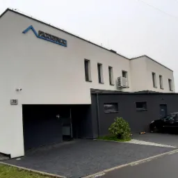 Nowoczesny budynek biurowy z białymi ścianami i ciemnymi elementami, logo firmy na elewacji, zaparkowany czarny samochód na wybrukowanym podjeździe.