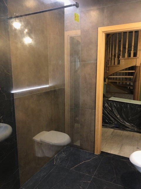 Wykończona łazienka z czarną marmurową podłogą, szarymi płytkami ściennymi i podświetlanym paskiem LED, widoczny fragment toalety i umywalki, w tle drewniane schody.