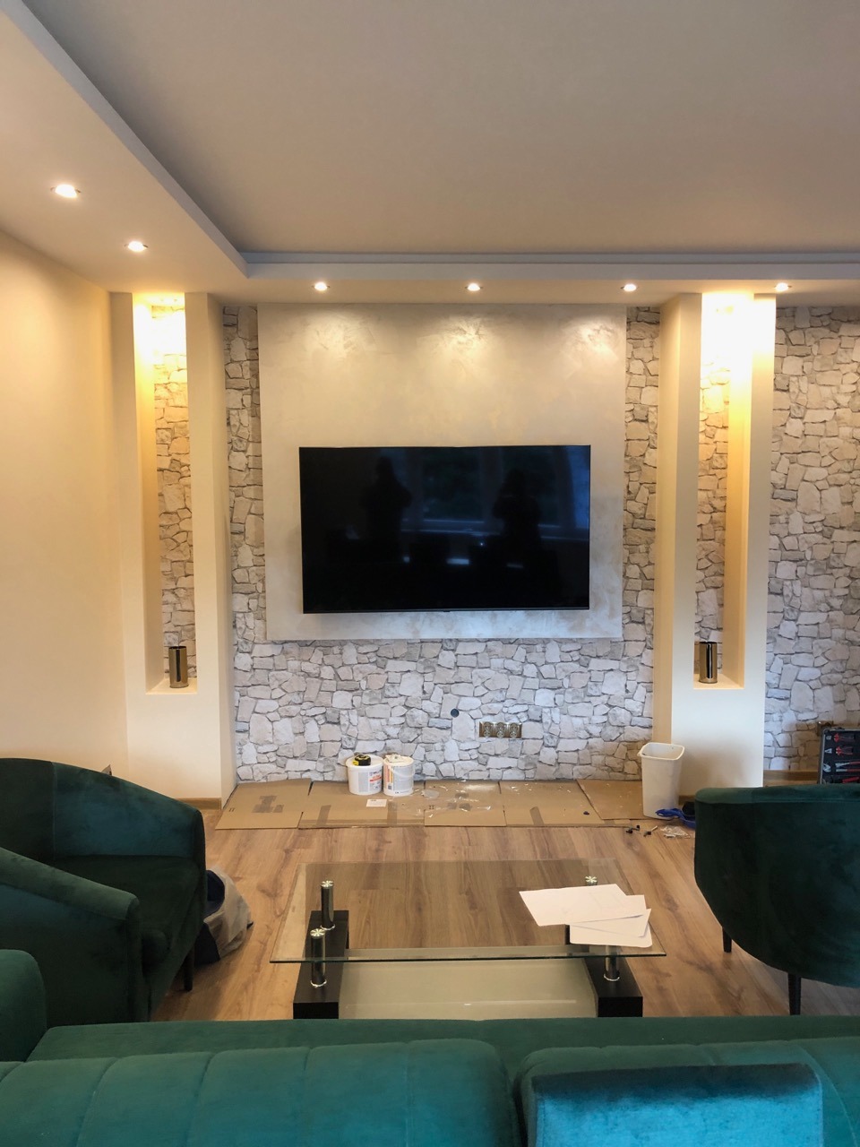 Salon w trakcie prac wykończeniowych: ściana z kamienia dekoracyjnego, zamontowany telewizor, widoczne puszki farb i zabezpieczenia podłogi, zielona sofa w pierwszym planie.