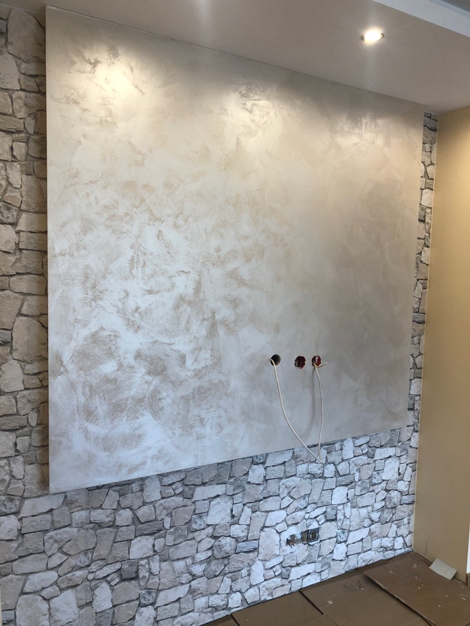 Ściana w pokoju z dekoracyjną, beżowo-szarą gładzią imitującą kamień, obok fragment ściany wykończony tapetą z motywem kamieni, widoczne gniazdka elektryczne i wystające przewody.