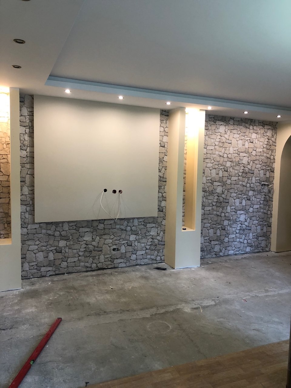 Pokój w trakcie prac wykończeniowych: ściana z imitacją kamienia, centralny panel przygotowany pod montaż telewizora z wystającymi kablami, surowa podłoga z poziomnicą w rogu.