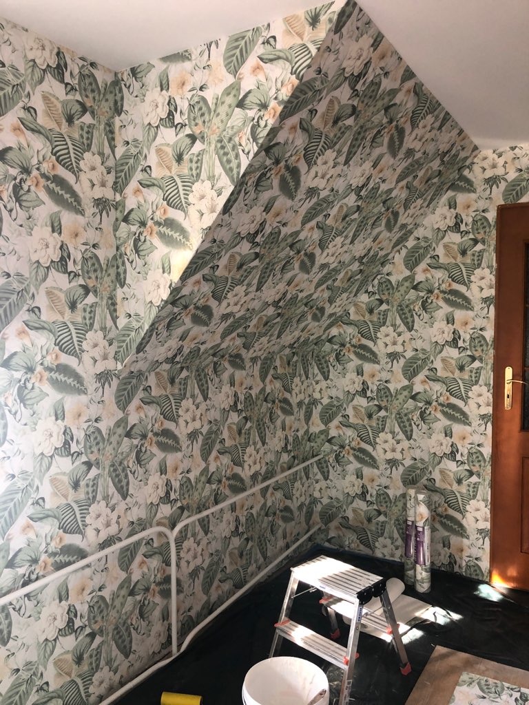 Pokój na poddaszu z ukończonym tapetowaniem ścian we wzór tropikalnych liści i kwiatów, widoczna drabina, wiadro z klejem i rolki tapety.