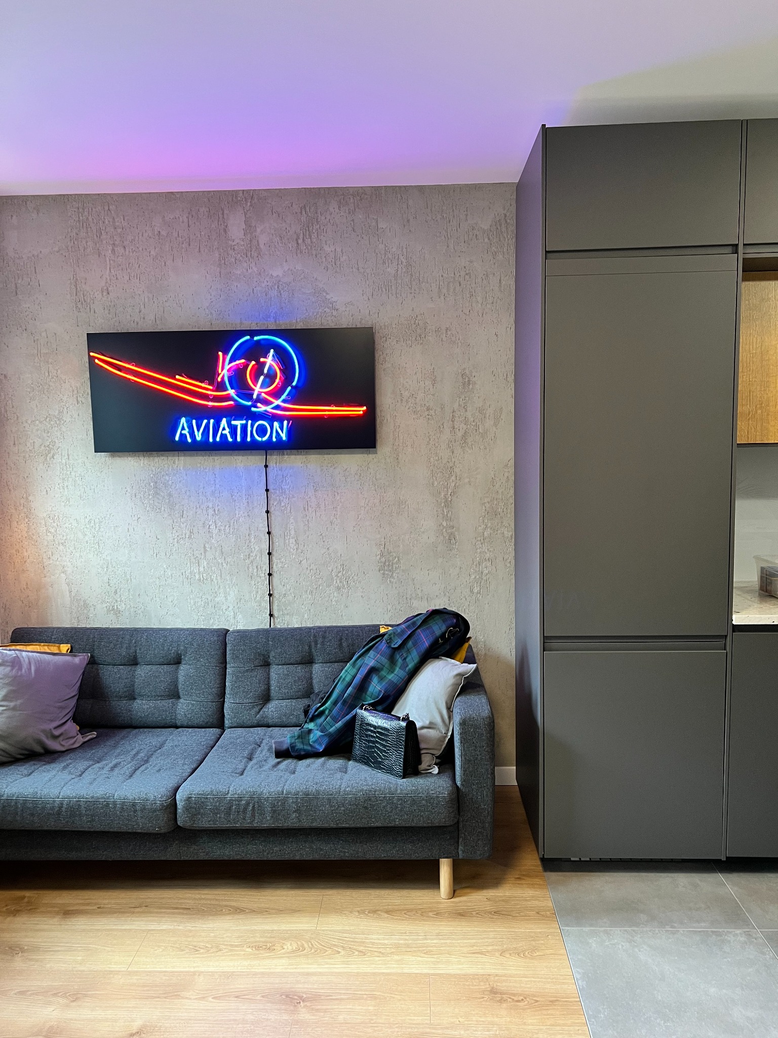 Szary narożnik z poduszkami i pledem w kratę, neonowy napis AVIATION na ścianie o fakturze betonu, wysoka zabudowa kuchenna w kolorze grafitowym, podłoga z paneli i płytek.
