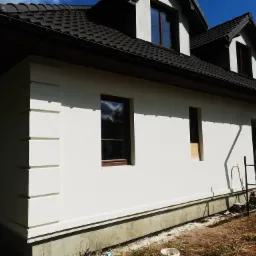 Jasny dom z ciemną dachówką, świeżo otynkowaną elewacją i dekoracyjnymi narożnikami, widoczny fragment rusztowania i niezagospodarowany teren wokół.