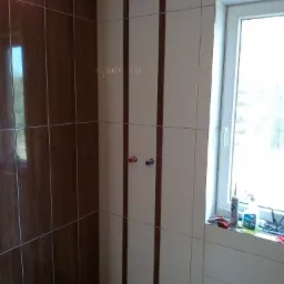 Wykończona ściana w łazience z brązowymi i białymi płytkami, widoczne wystające rury instalacji wodnej oraz okno z akcesoriami do montażu.