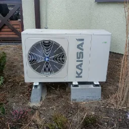 Zewnętrzna jednostka pompy ciepła Kaisai, umieszczona na betonowych bloczkach przy ścianie domu z dekoracyjną elewacją z kamienia, otoczona roślinnością.