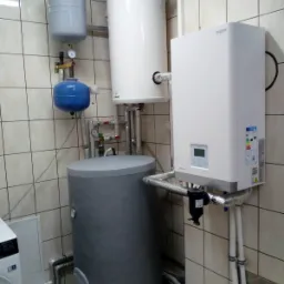 Kompaktowa kotłownia: szary zasobnik ciepłej wody użytkowej, biały bojler oraz wiszący piec grzewczy z wyświetlaczem, połączone rurami w pomieszczeniu wyłożonym kremowymi płytkami.