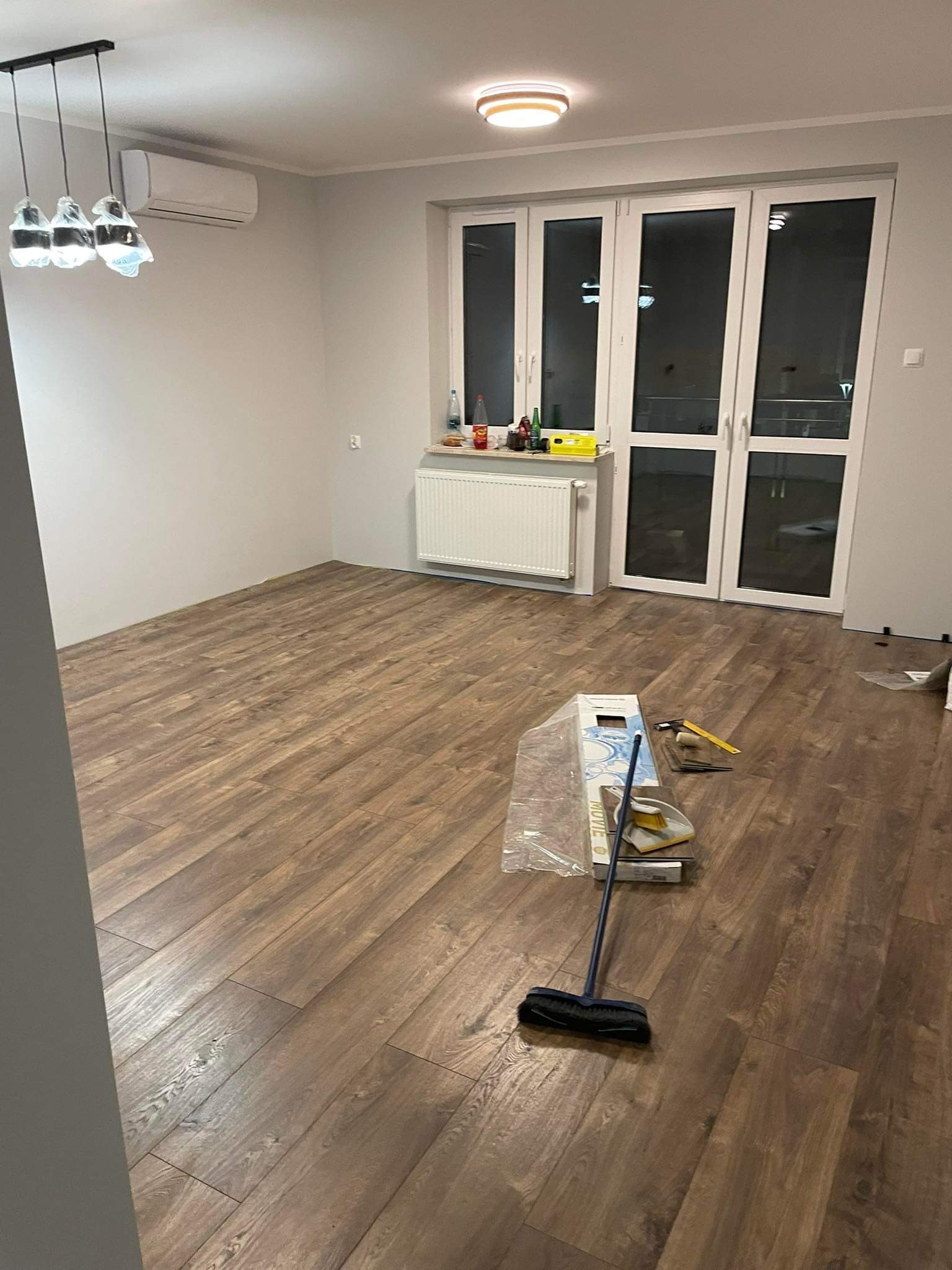 Pokój z nowo ułożoną, brązową podłogą z paneli imitujących drewno, widoczne narzędzia do montażu i szczotka do zamiatania, okno balkonowe z widokiem na noc.