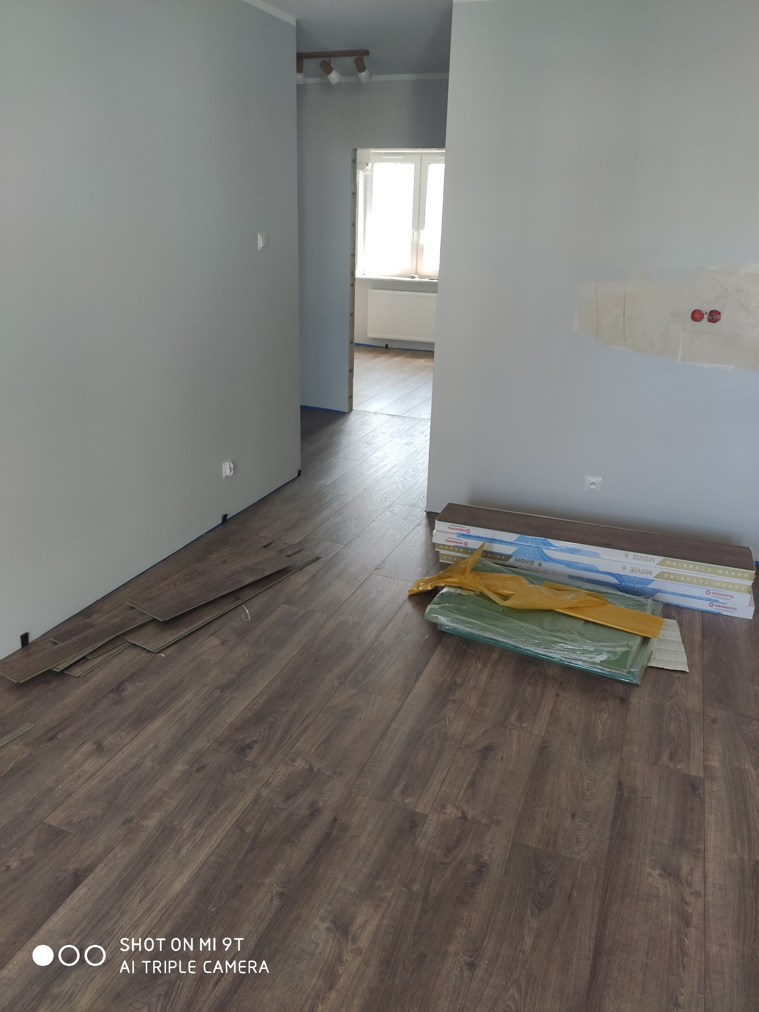 Układanie paneli podłogowych w trakcie remontu: widoczne panele o ciemnym odcieniu drewna, częściowo ułożone przy ścianie, obok paczki nowych paneli w opakowaniu i folii ochronnej, na tle szarych...