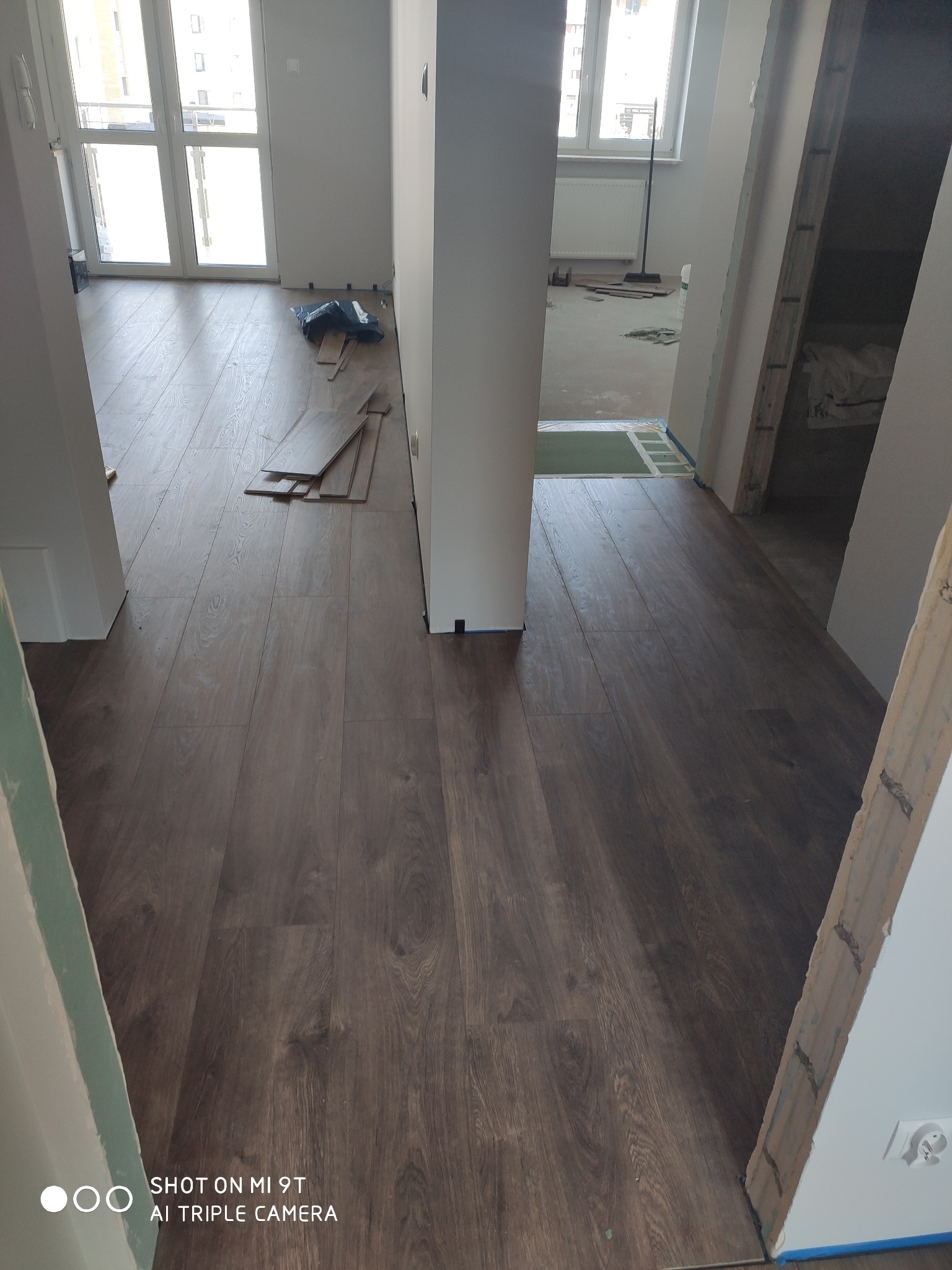 Wnętrze w trakcie wykańczania: ułożona podłoga z paneli w odcieniach brązu, widoczne przygotowane panele do ułożenia w rogu pomieszczenia, surowe wykończenie ościeżnic drzwiowych.