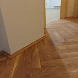 Ułożona drewniana podłoga w jodełkę, widoczna ściana z listwą przypodłogową oraz fragment drzwi do innego pomieszczenia z grzejnikiem.