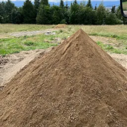 Kupa ziemi w kształcie piramidy na przygotowanym terenie budowy, w tle drzewa i zarys gór na horyzoncie.