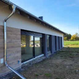 Fragment elewacji domu jednorodzinnego z drewnopodobną okładziną, widoczne rynny, okna z roletami zewnętrznymi i fragment niezabezpieczonego terenu wokół budynku.