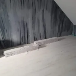 Poddasze z nowymi panelami podłogowymi Kronopol Room w kolorze jasnego drewna, gotowymi do ułożenia, ściany pomalowane w szaro-czarne, nieregularne wzory, widoczny fragment wiklinowego fotela.