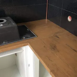 Fragment nowej kuchni z drewnianym blatem, czarnymi mozaikowymi płytkami i białymi szafkami, widoczny fragment zlewu.