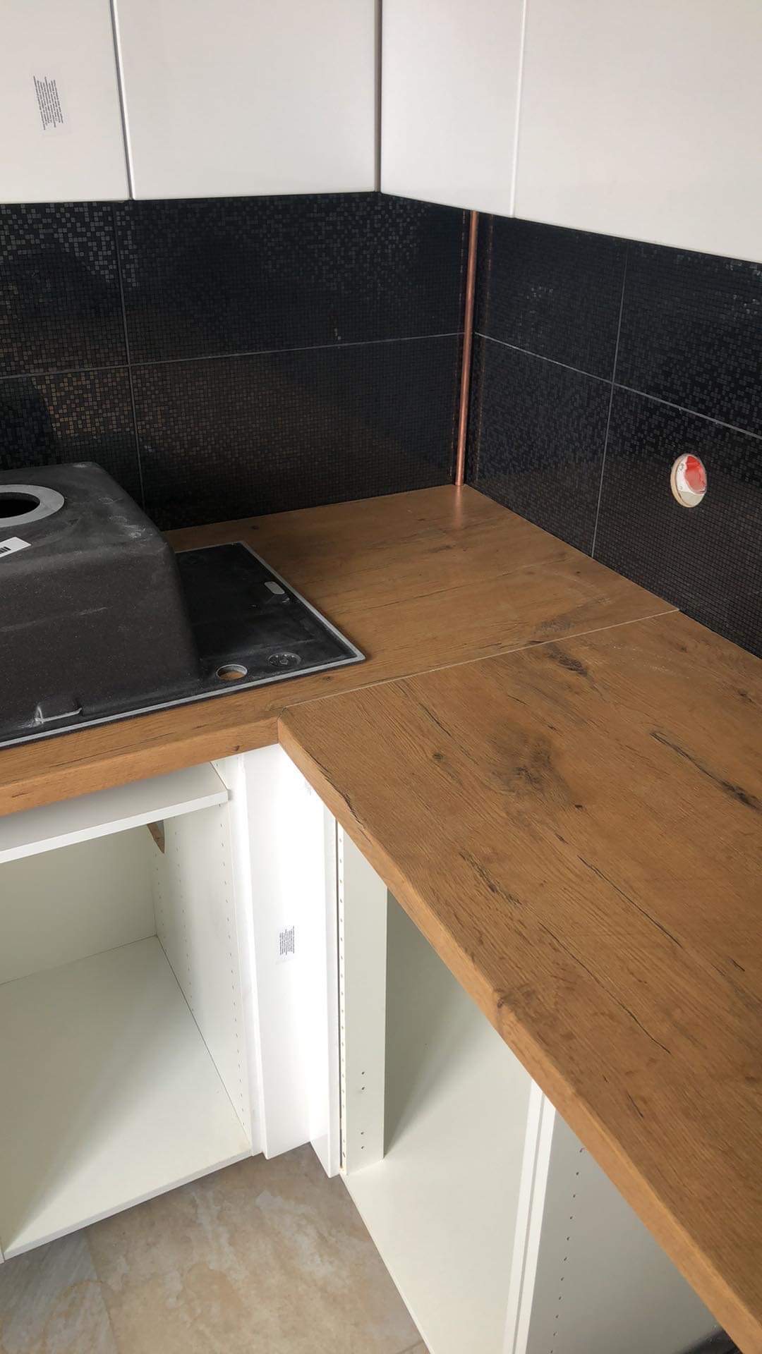 Fragment nowej kuchni z drewnianym blatem, czarnymi mozaikowymi płytkami i białymi szafkami, widoczny fragment zlewu.