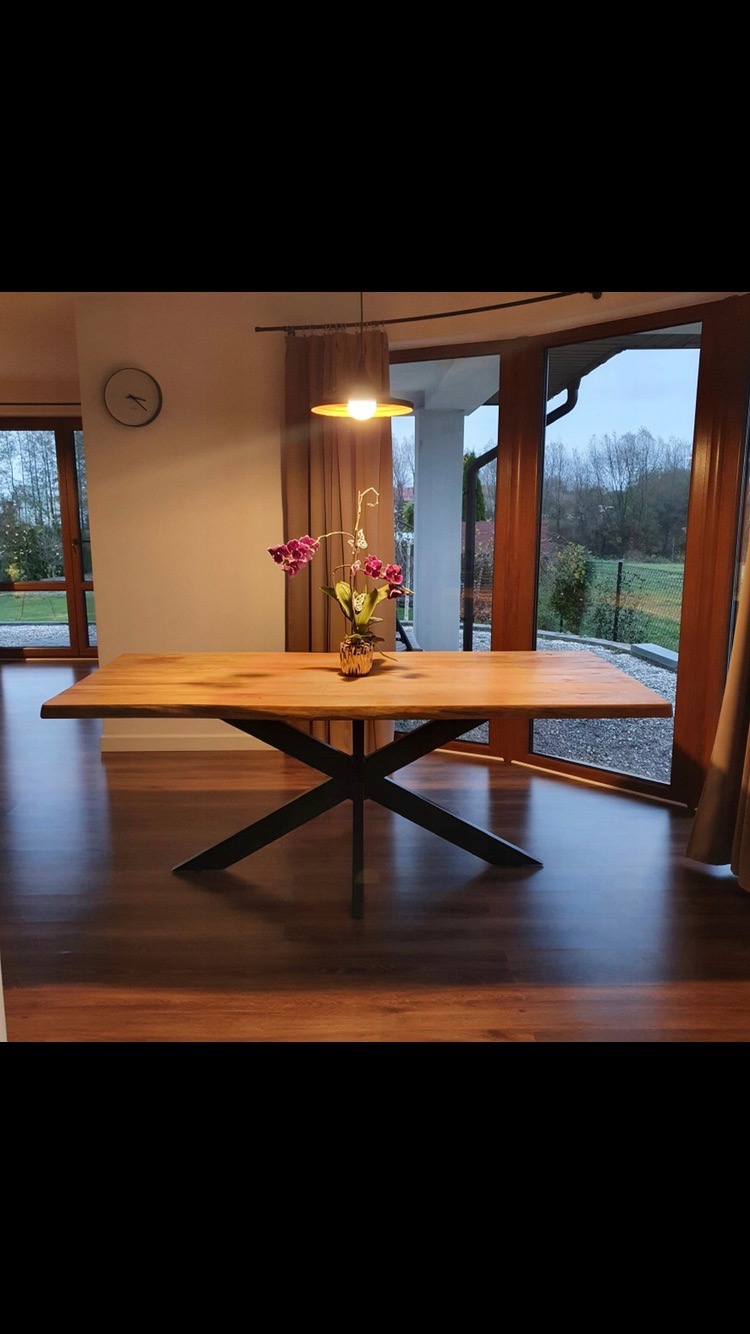 Stół z blatem z jasnego drewna i czarną, krzyżową podstawą, na nim wazon z fioletowymi orchideami, w tle okna z widokiem na ogród.