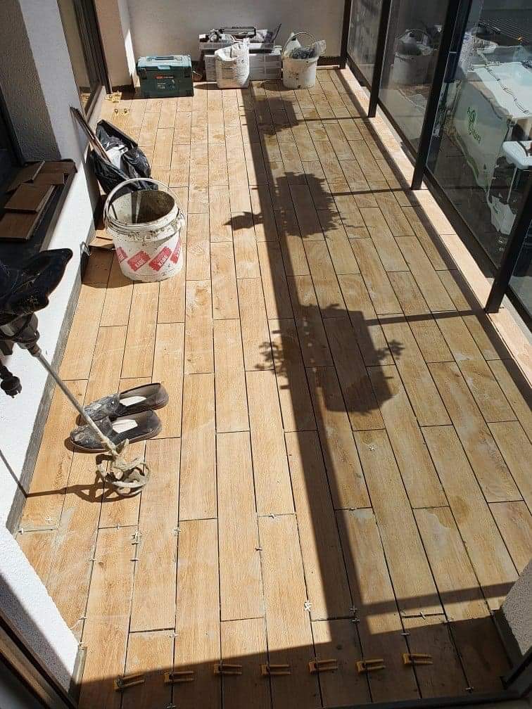 Układanie podłogi z płytek imitujących drewno na balkonie, widoczne narzędzia, wiadro i krzyżyki dystansowe, częściowo ukończona praca.
