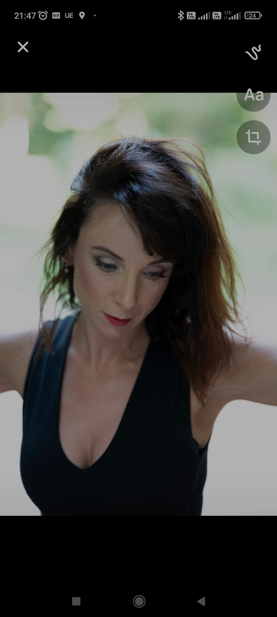 Portret kobiety z delikatnym makijażem oczu i ust w odcieniu czerwieni, patrzącej w dół, z rozpuszczonymi ciemnymi włosami i czarną sukienką na rozmytym zielonym tle.