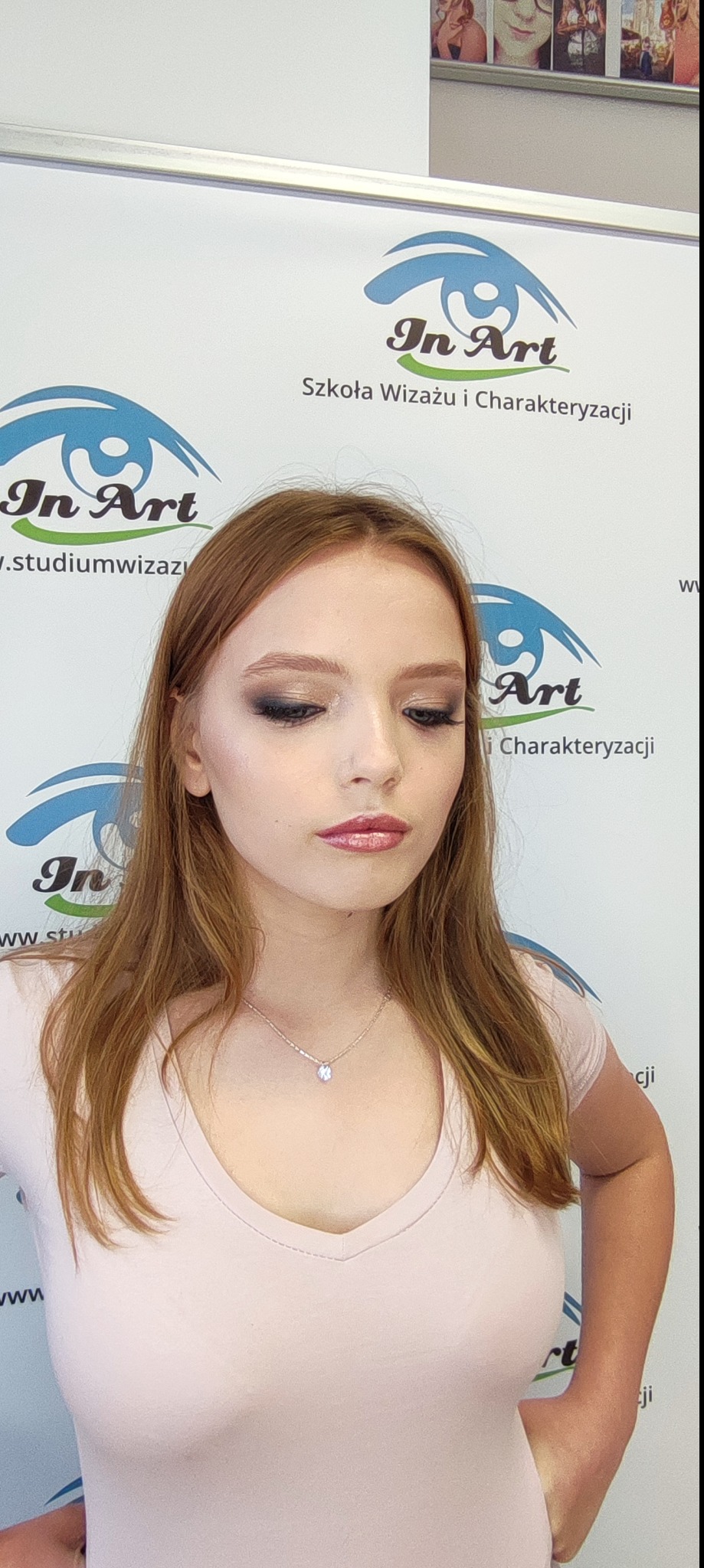 Portret młodej kobiety z profesjonalnym makijażem oczu w odcieniach szarości i błyszczącymi ustami w odcieniu różu, na tle roll-upu szkoły wizażu.