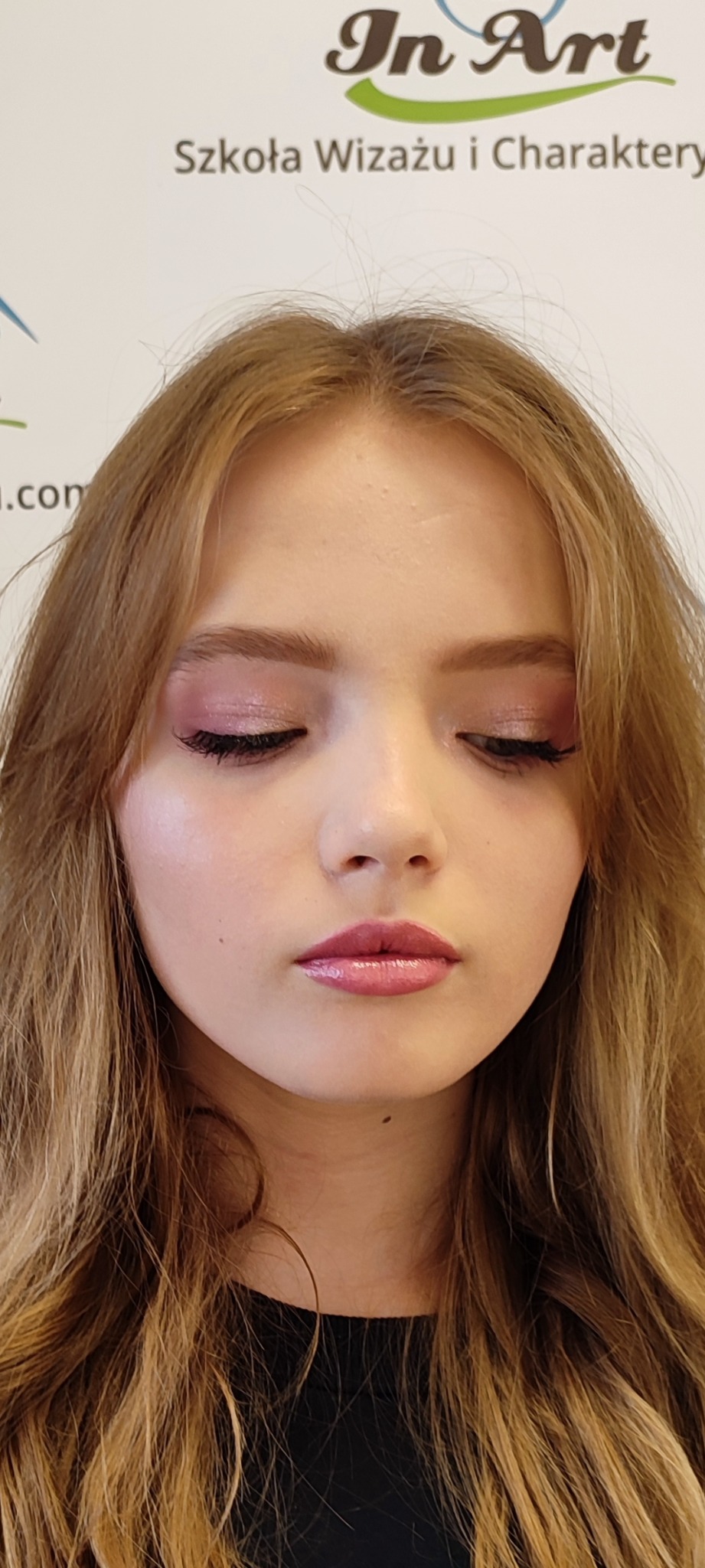 Portret młodej kobiety z delikatnym, różowym makijażem oczu i ust. Widoczne logo szkoły wizażu w tle.