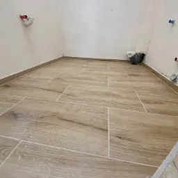 Ułożona podłoga z płytek imitujących drewno w jasnym odcieniu, widoczne fragmenty ścian z instalacjami, małe okno w górnej części ściany, wiadro z narzędziami w rogu pomieszczenia.