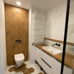 Nowoczesna łazienka z drewnianymi akcentami: zabudowa toalety z płyty imitującej drewno, heksagonalne płytki podłogowe w odcieniach bieli i drewna, biała szafka pod umywalkę z drewnianym blatem...