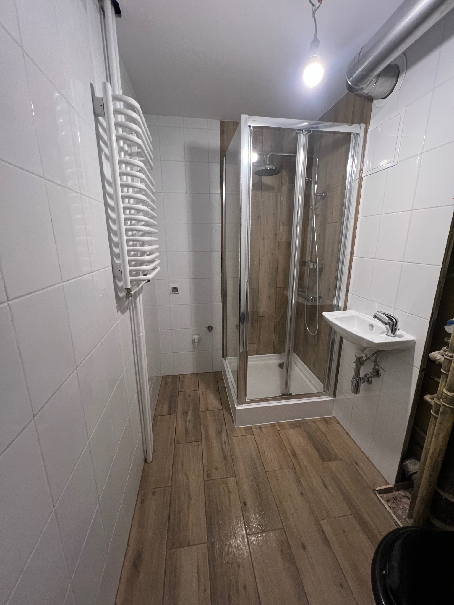 Ukończona łazienka z kabiną prysznicową z chromowanymi ramami, małą umywalką narożną i grzejnikiem drabinkowym na białych kafelkach ściennych oraz podłogą imitującą drewno.